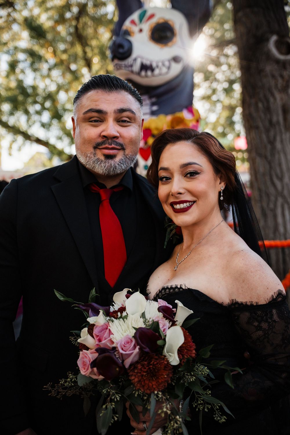 A couple in black wedding attire poses outdoors with a gothic-themed floral bouquet featuring dark and light blooms.
