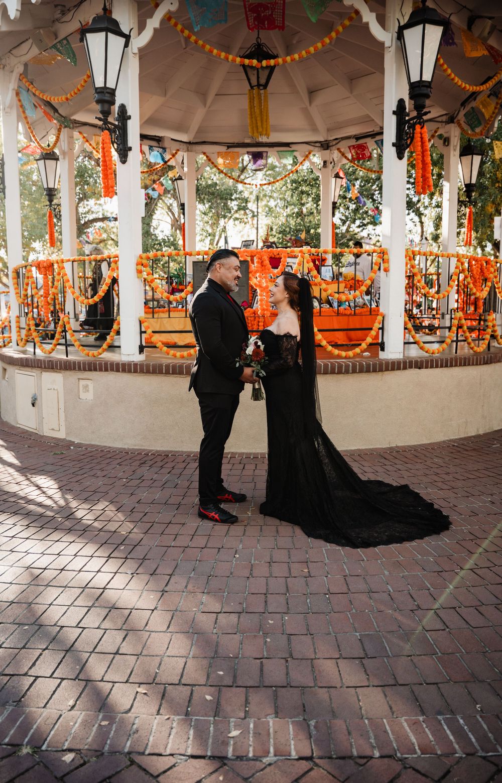 A gothic wedding couple poses under an orange-decorated pavilion with brick flooring.