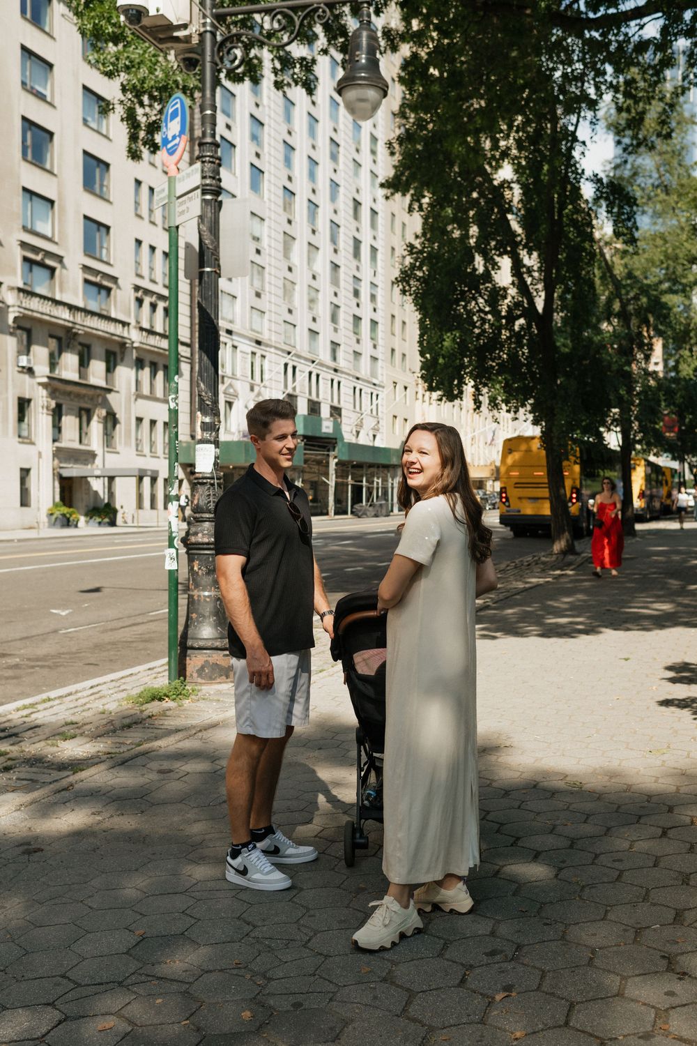 Two people interact on a city sidewalk with tall apartment buildings and trees in the background.