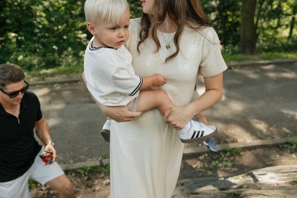 Family wearing coordinated white outfits embraces during outdoor photo session in a sunlit wooded park setting.