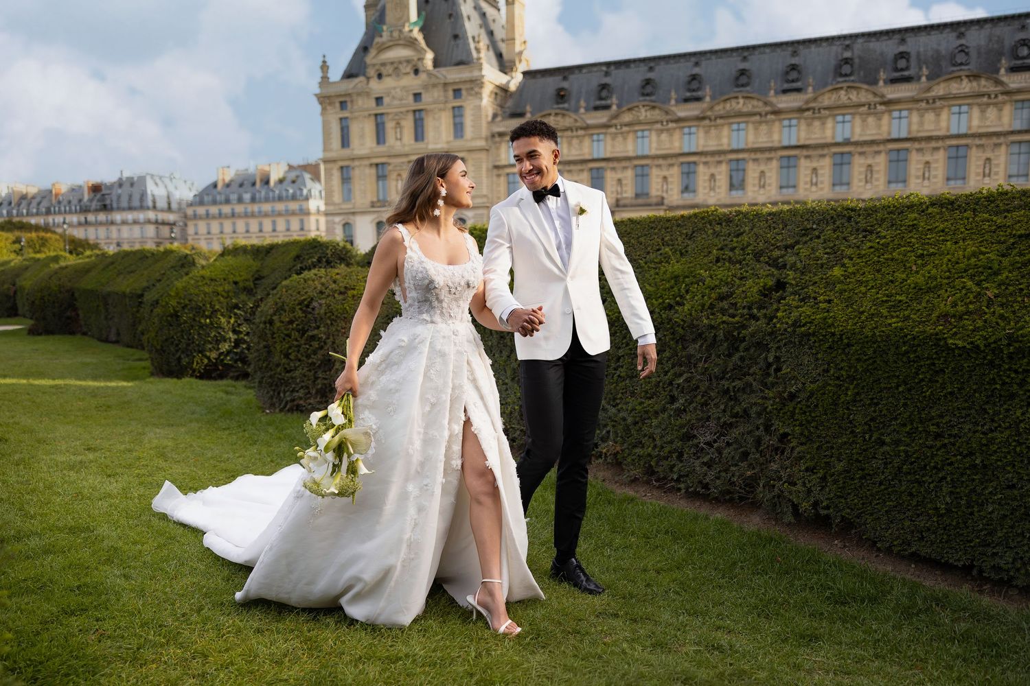 Elegant wedding couple poses in front of a grand historic palace with manicured gardens.