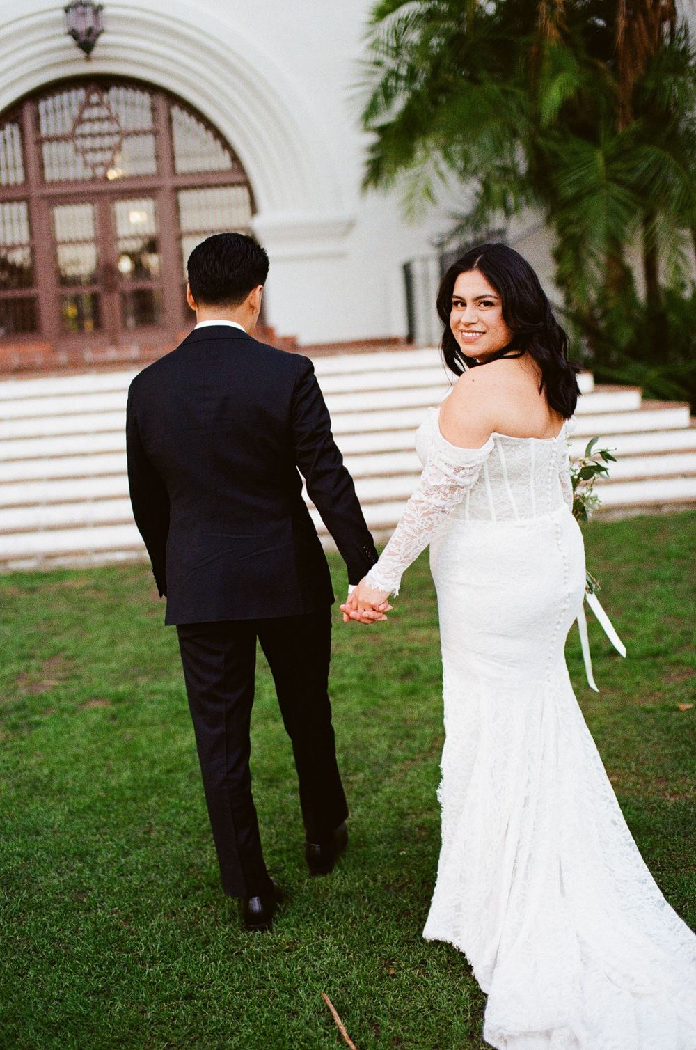 35mm film photo of a bride in a fitted white lace gown looks back while holding hands with her groom on a grassy lawn by an arched entrance.