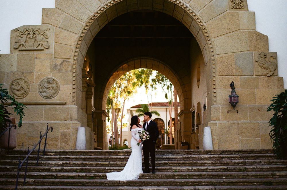 A couple stands beneath a grand stone archway with ornate carvings at a Mediterranean-style venue.