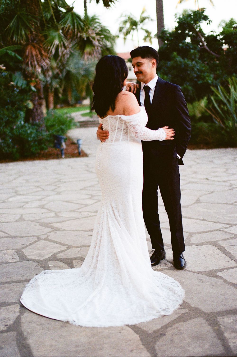 A romantic wedding moment captured on a stone pathway surrounded by tropical palm trees and lush greenery.