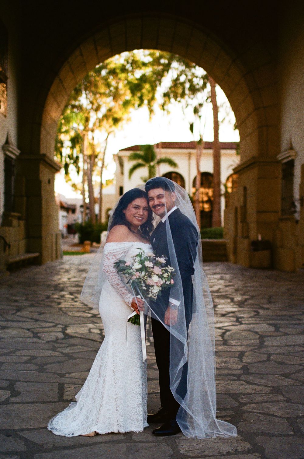 A wedding couple stands under a stone archway on a cobblestone path at sunset, with flowing veil and floral bouquet.