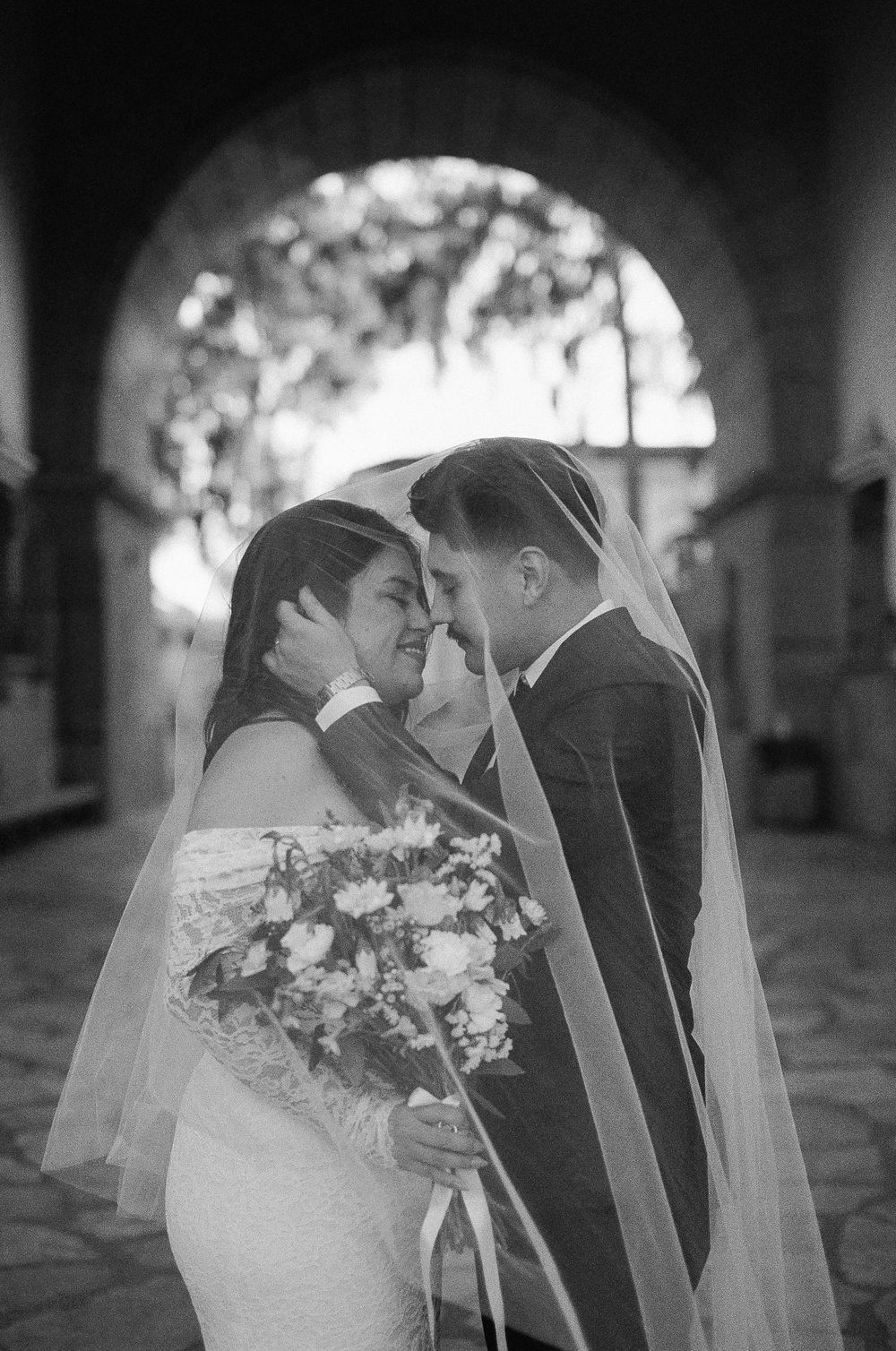 Black and white photo of newlyweds sharing an intimate moment under an archway with a bridal bouquet and veil at Santa Barbara Courthouse.