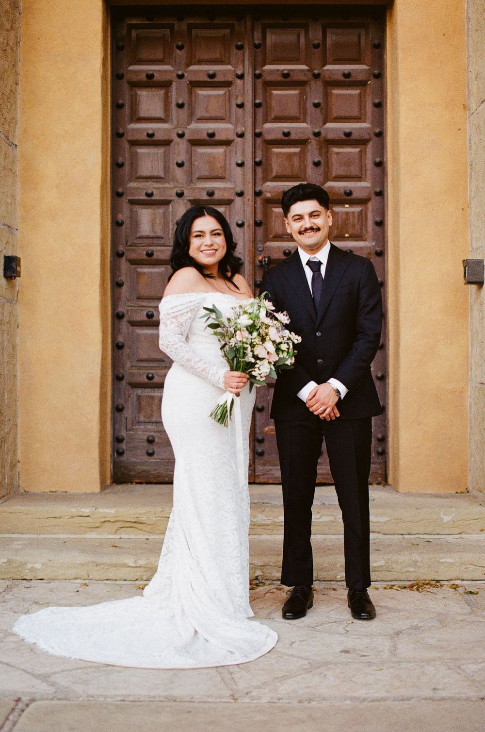 A lovely wedding couple poses in front of a large wooden door at a historic building.