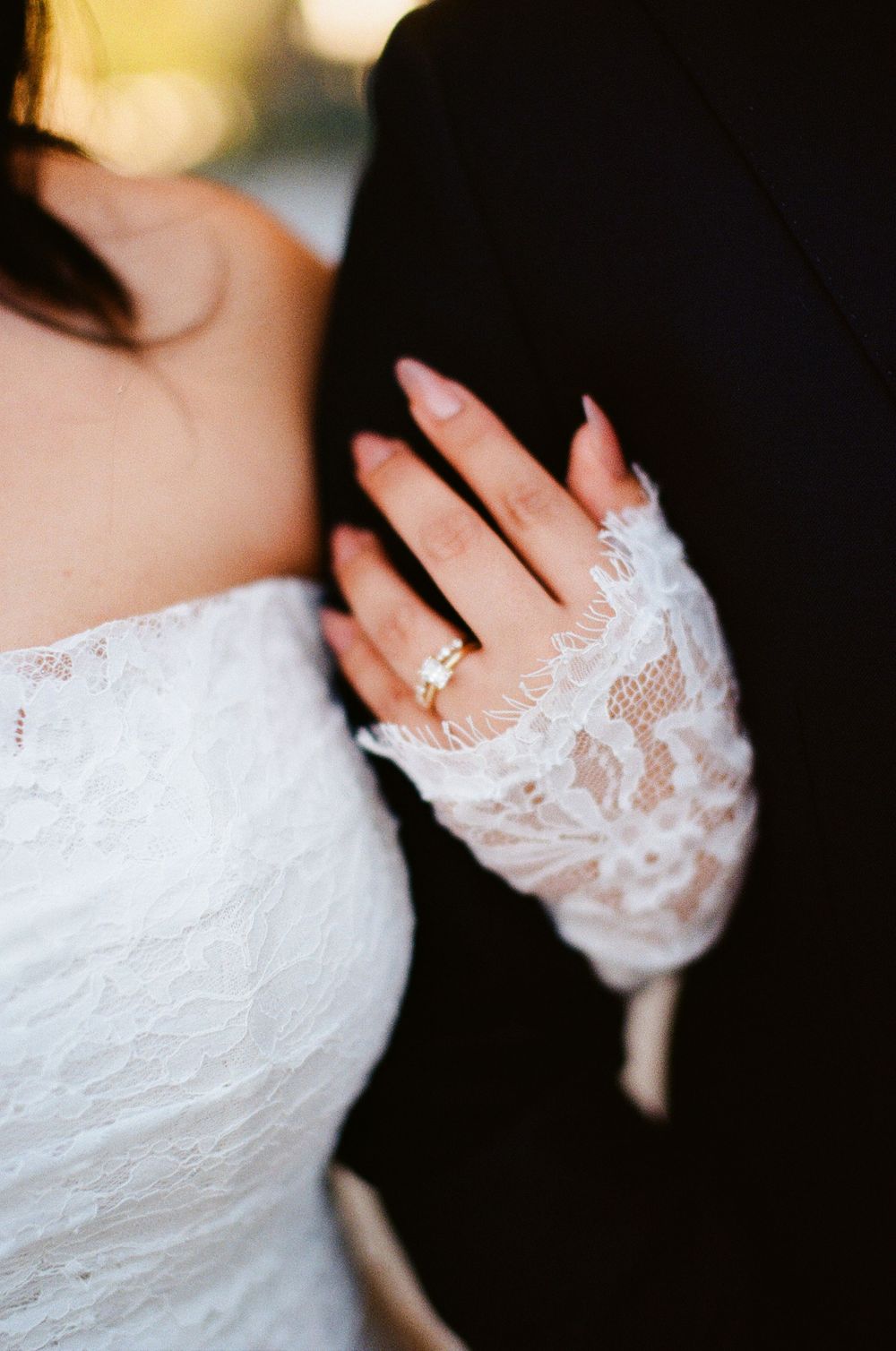 Close-up of a bride's hand with engagement ring wearing delicate lace sleeve details against a black suit.