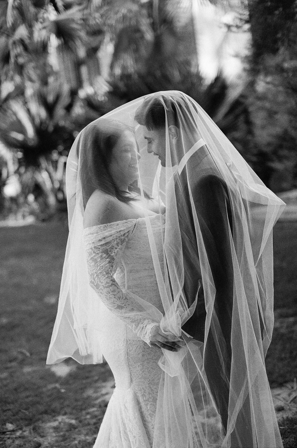 A romantic black and white wedding photo showing a couple sharing an intimate moment under a flowing veil outdoors.