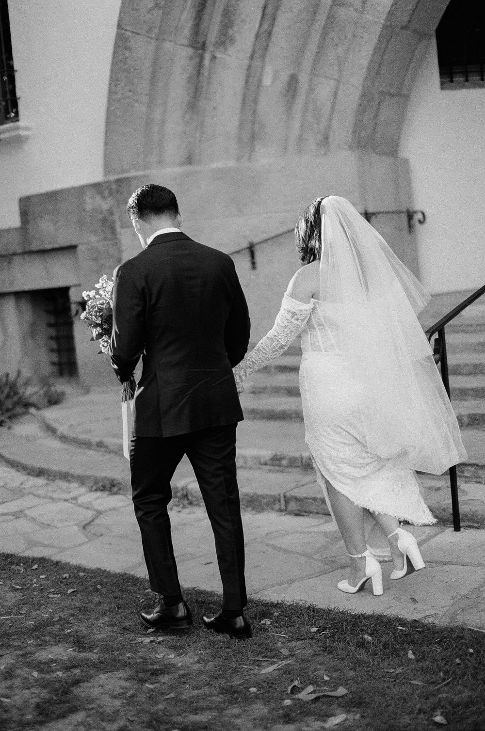 A bride in white dress and veil walks up stone church steps alongside a groom in black suit during a wedding ceremony.