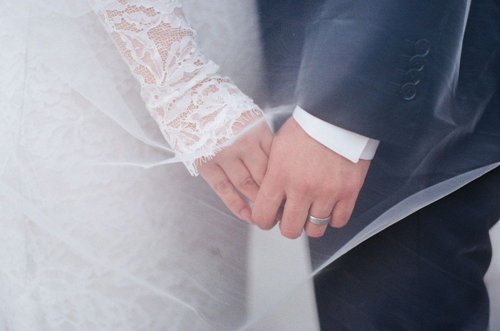 A close-up of newlyweds holding hands, showing white lace from the bride's sleeve and dark suit jacket of the groom.