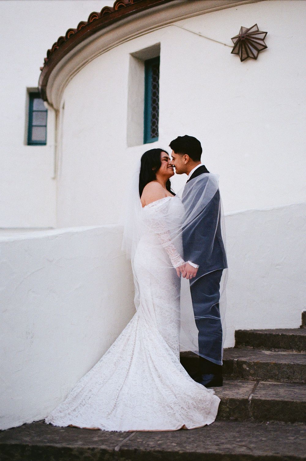 35mm film photo of a romantic wedding couple stands against a white stucco wall, sharing an intimate moment on stone steps.