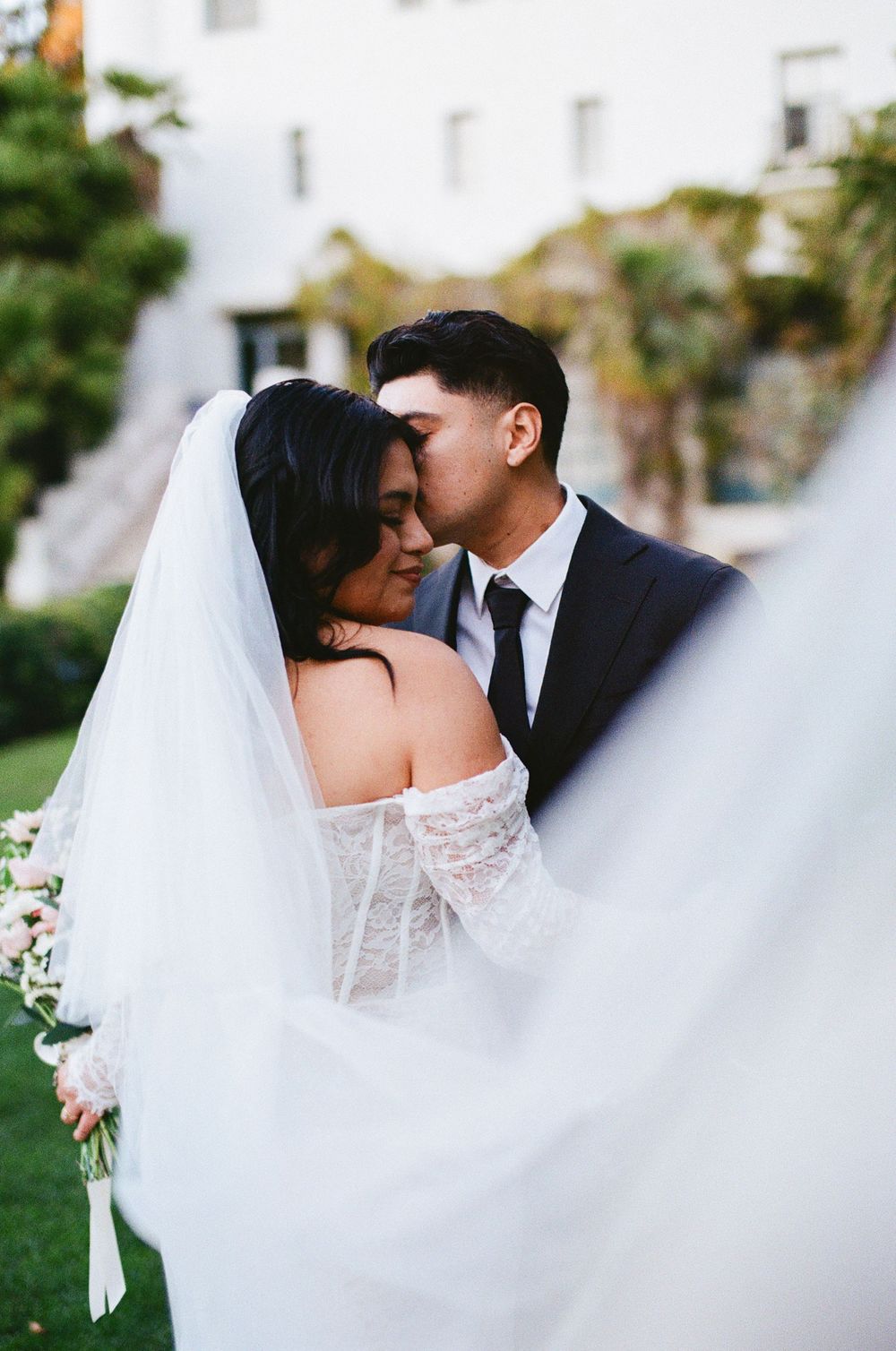 A tender wedding moment captured with flowing white veil and lace dress against a garden backdrop at the Santa Barbara Courthouse.