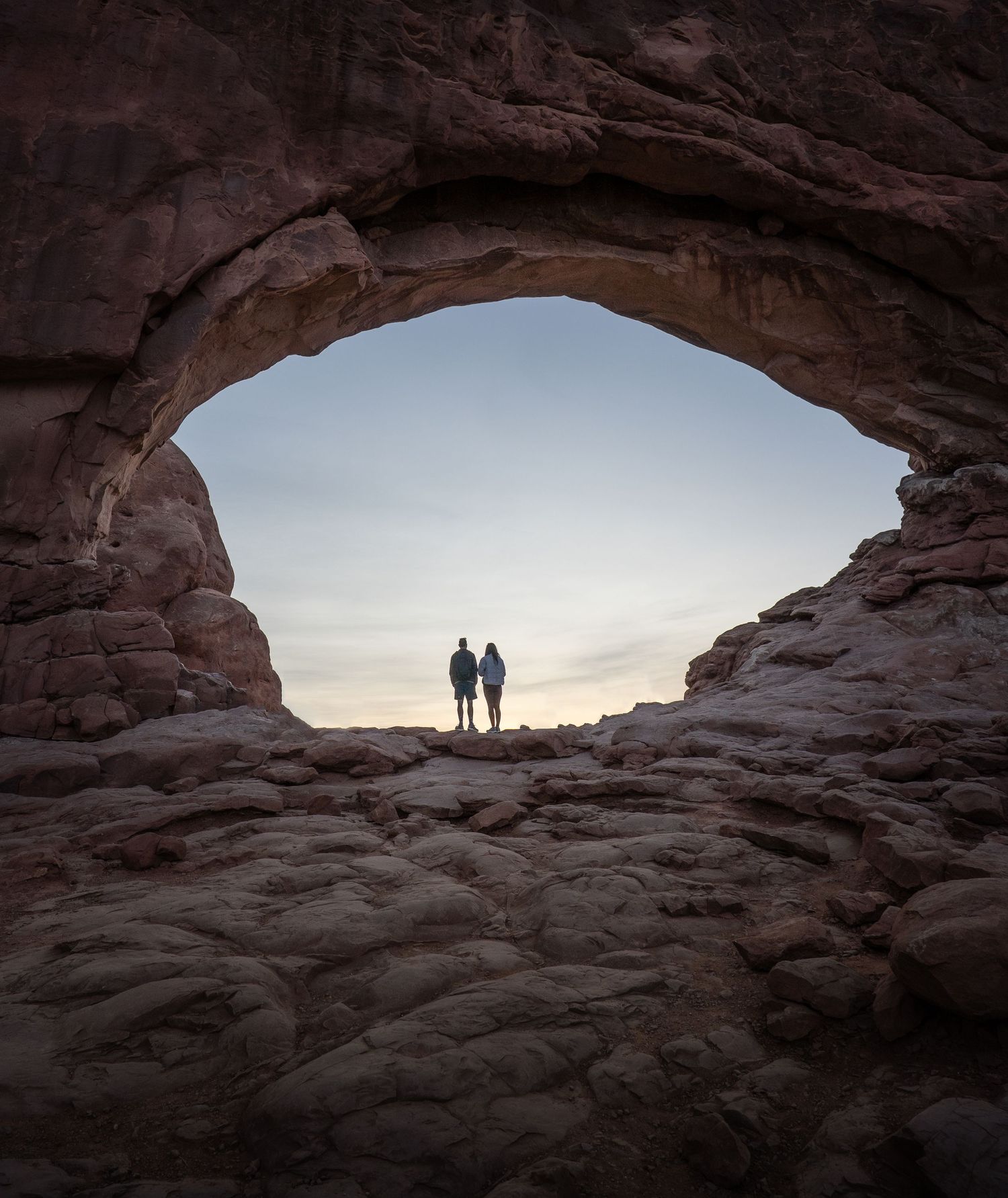 Two silhouettes stand framed within a natural rock arch formation at sunset in a desert landscape in Arches National Park in Moab, Utah.