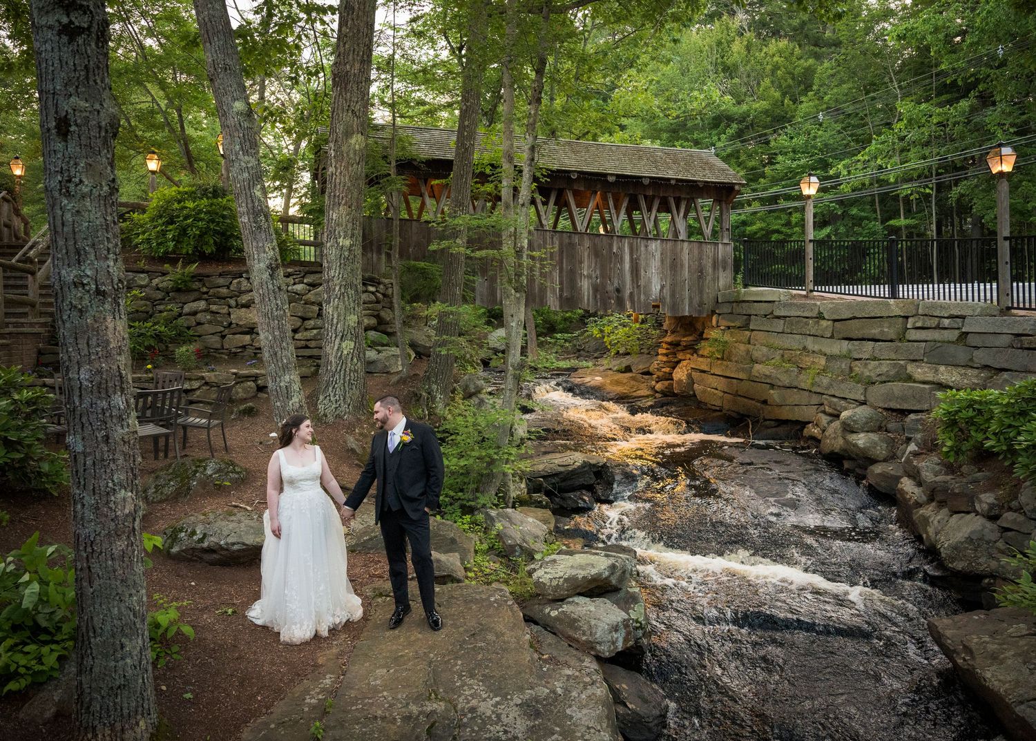 A couple stands together on a stone ledge in a wooded outdoor setting during a wedding photoshoot.