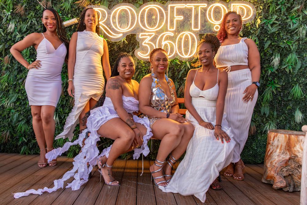 A group in coordinated white outfits poses together at an outdoor rooftop venue with illuminated signage.