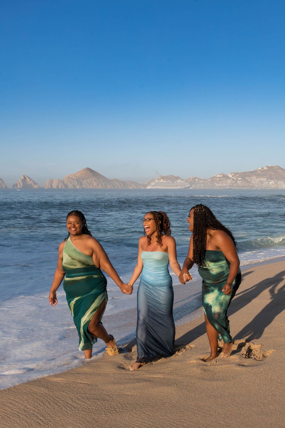 Three friends in flowing dresses walk hand in hand along a scenic beach with mountains in the background.