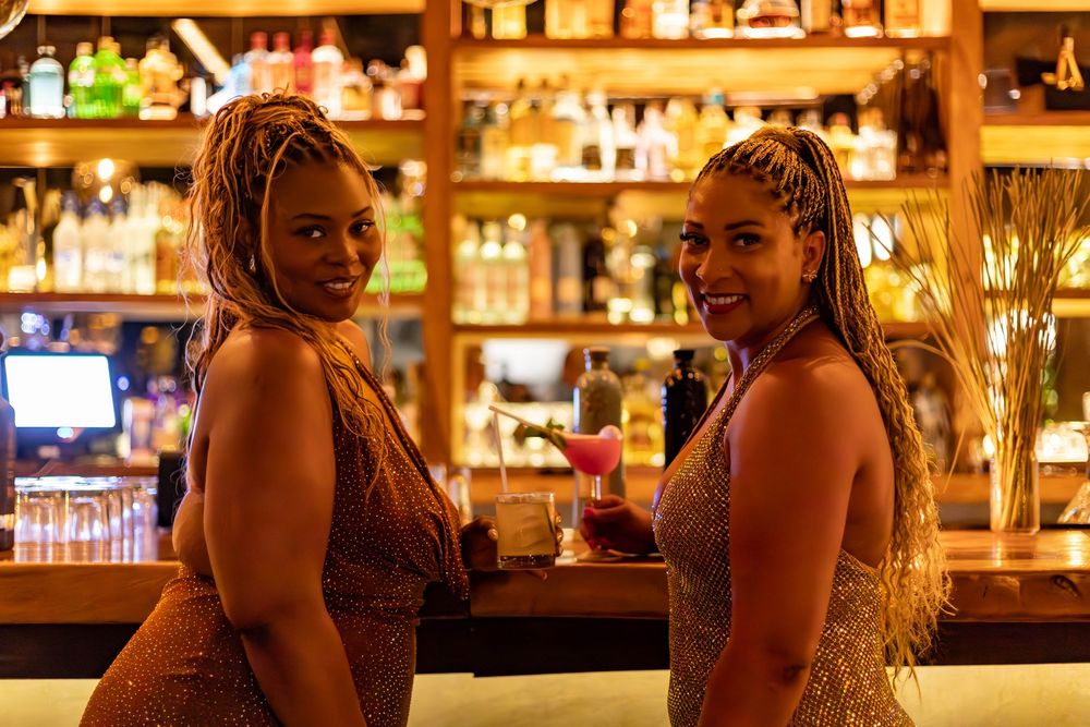 Two people sit at a warmly lit bar counter with bottles and glassware visible in the background.