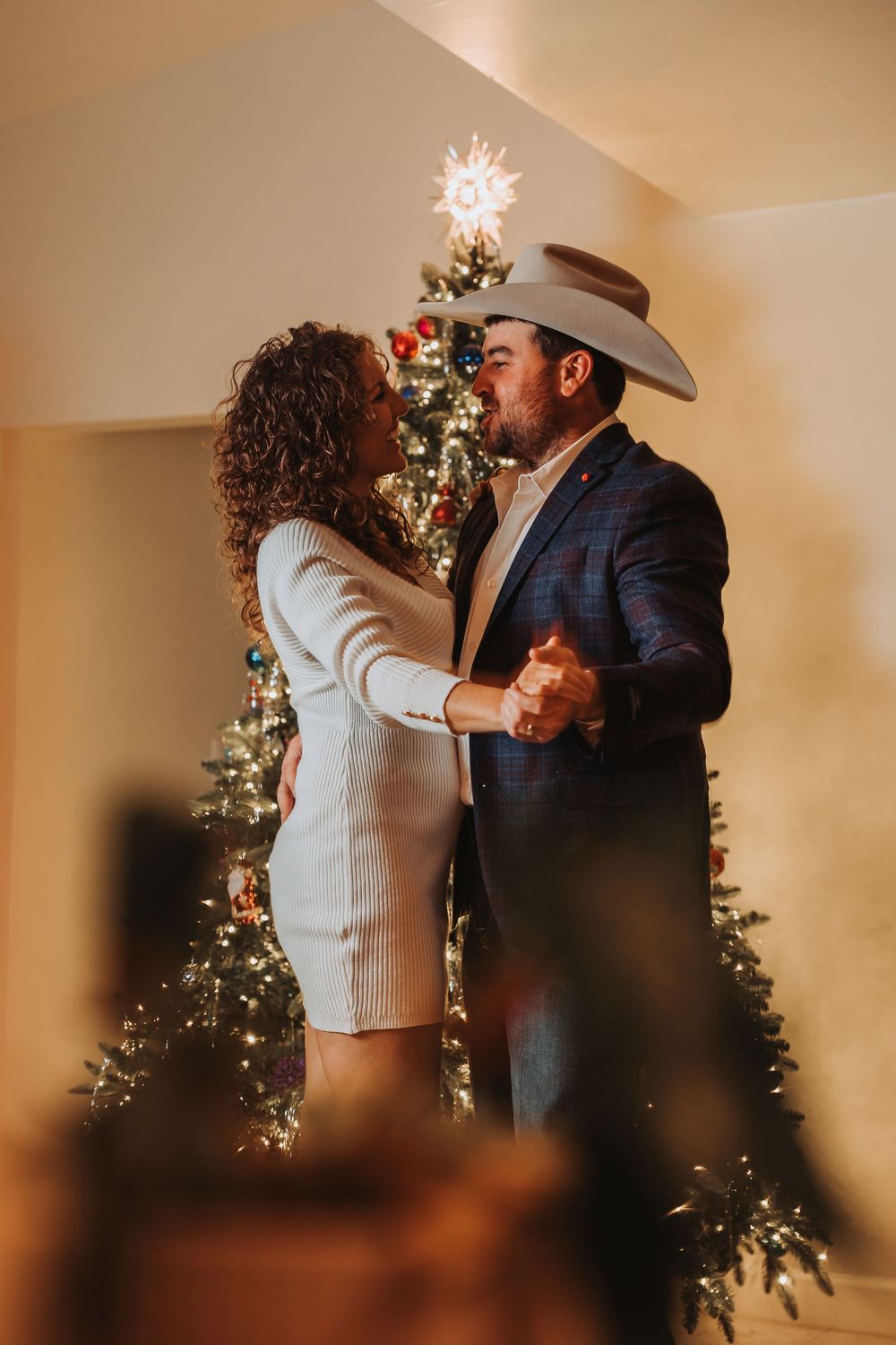 A couple shares a romantic dance moment in front of a decorated Christmas tree in a warm-lit room.