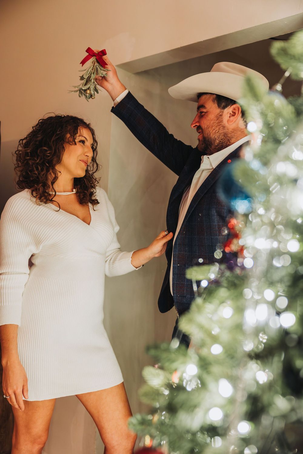 A playful couple dances together in front of a Christmas tree with twinkling lights and decorations in the background.