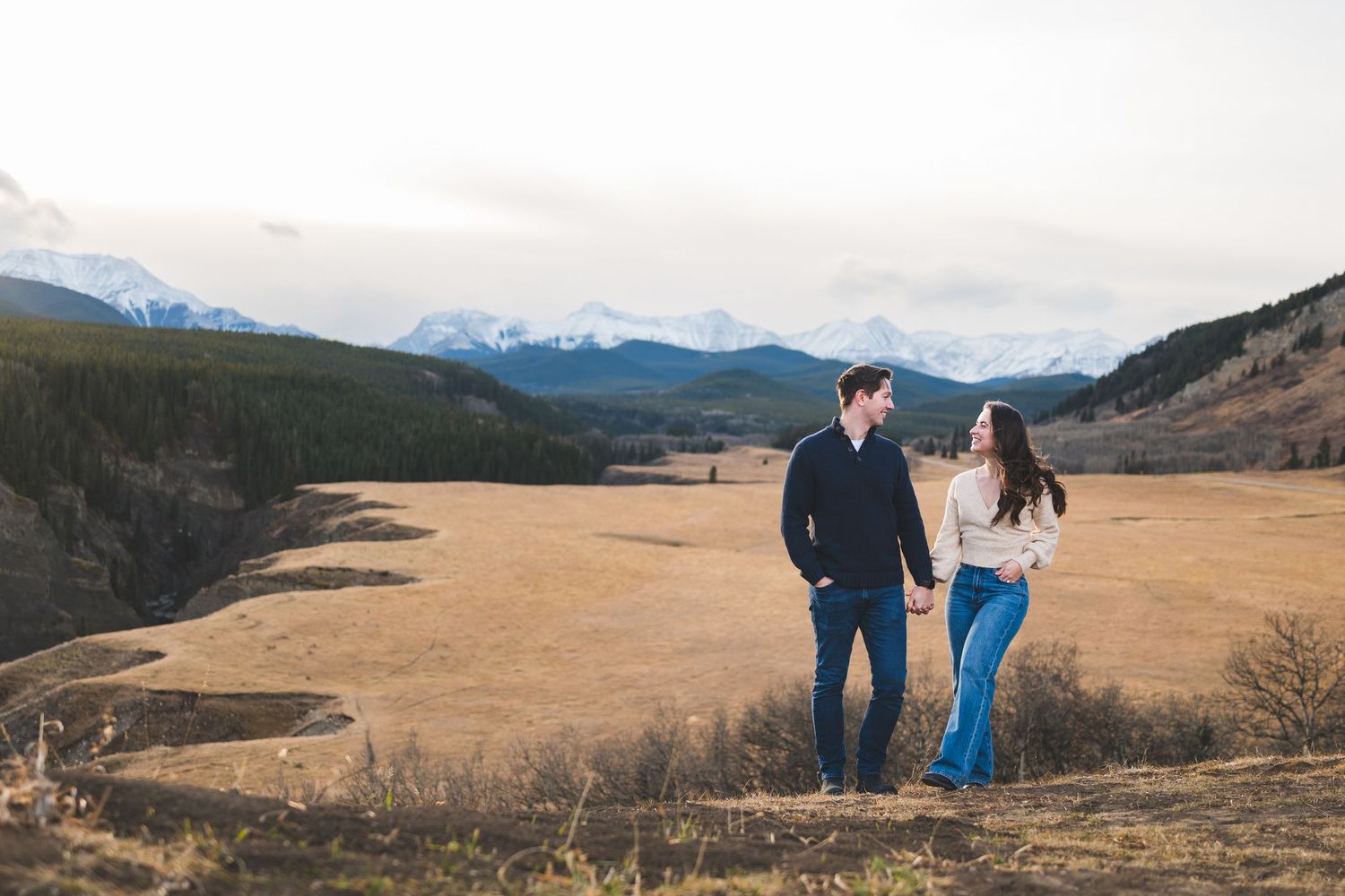 Couple walking together across a scenic mountain meadow with distant peaks in the background.