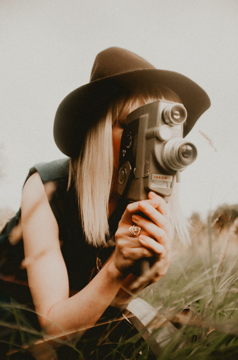 Photographer in a brown hat using a vintage movie camera in a field during golden hour.