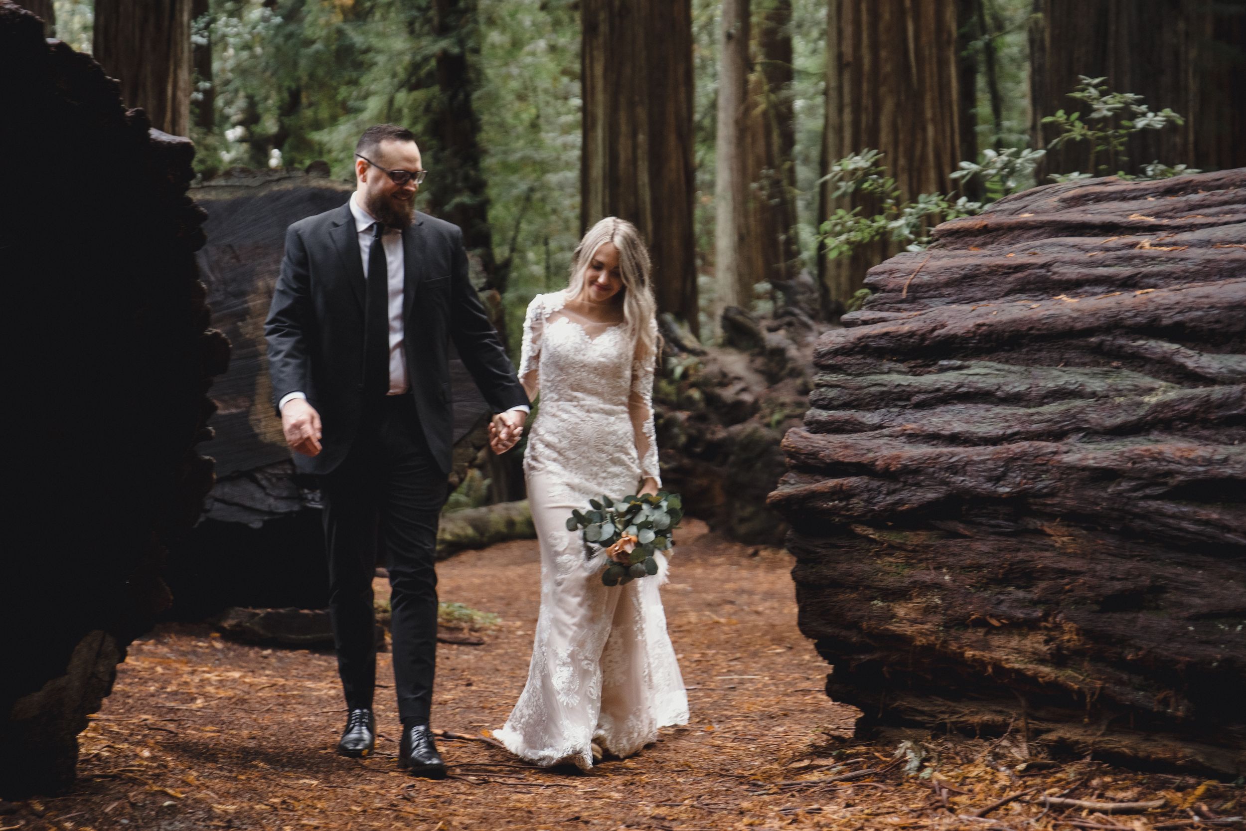 Bride and groom walk hand-in-hand through a lush redwood forest, she in a lace gown holding a bouquet.