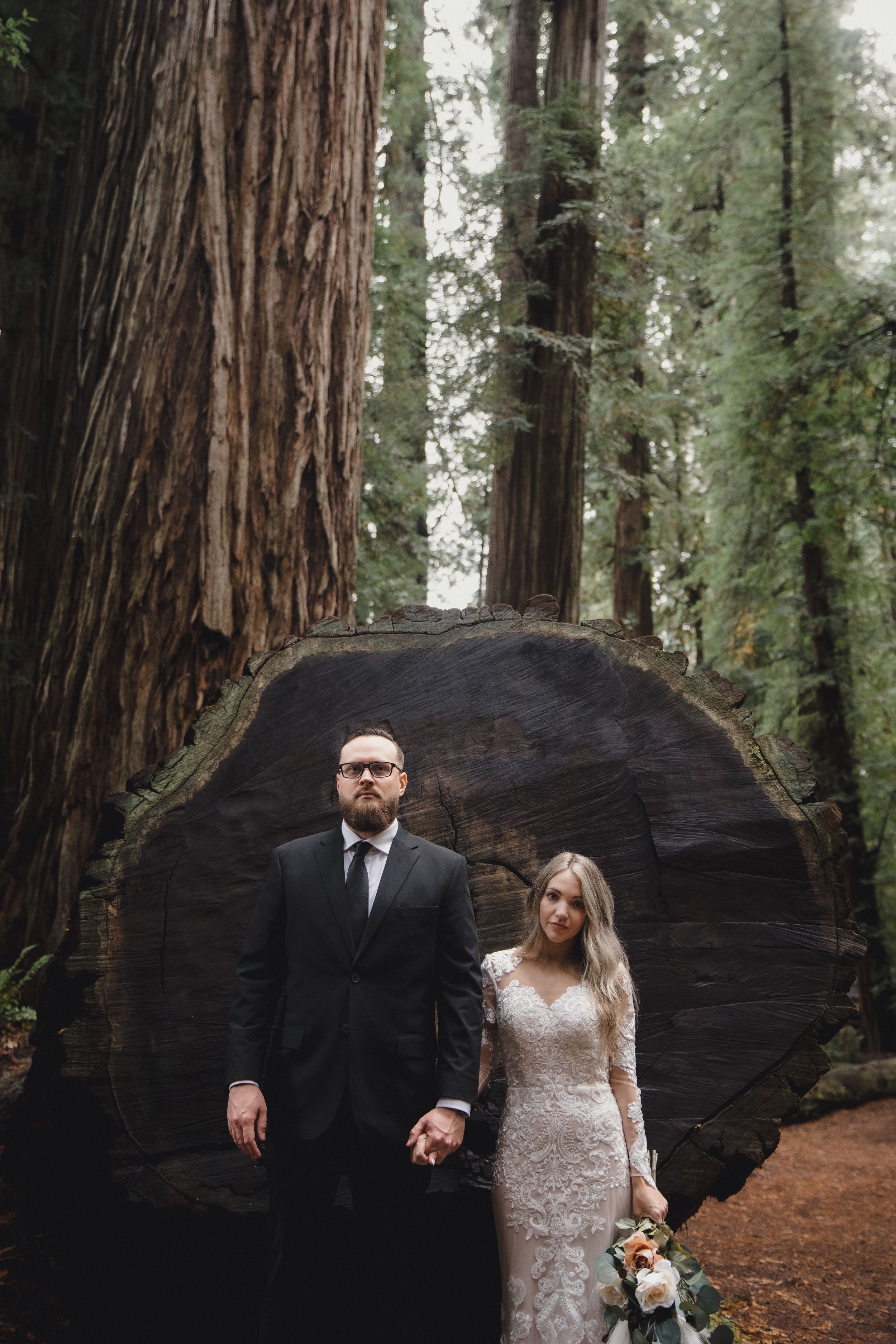 Bride and groom hold hands before a massive redwood stump in a lush forest wedding portrait.
