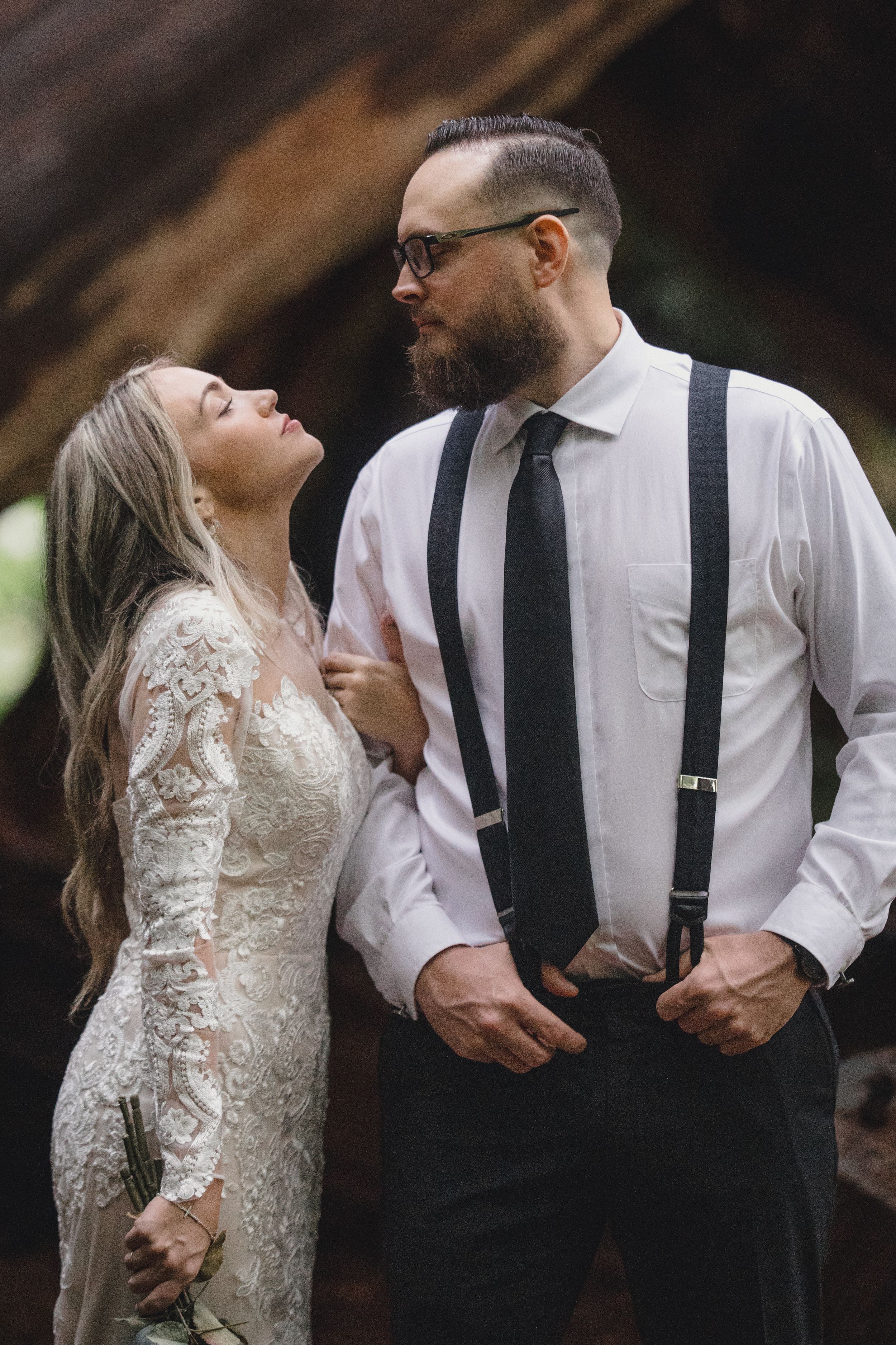 Bride in lace gown gazes up at bearded groom in white shirt with suspenders during romantic outdoor wedding.