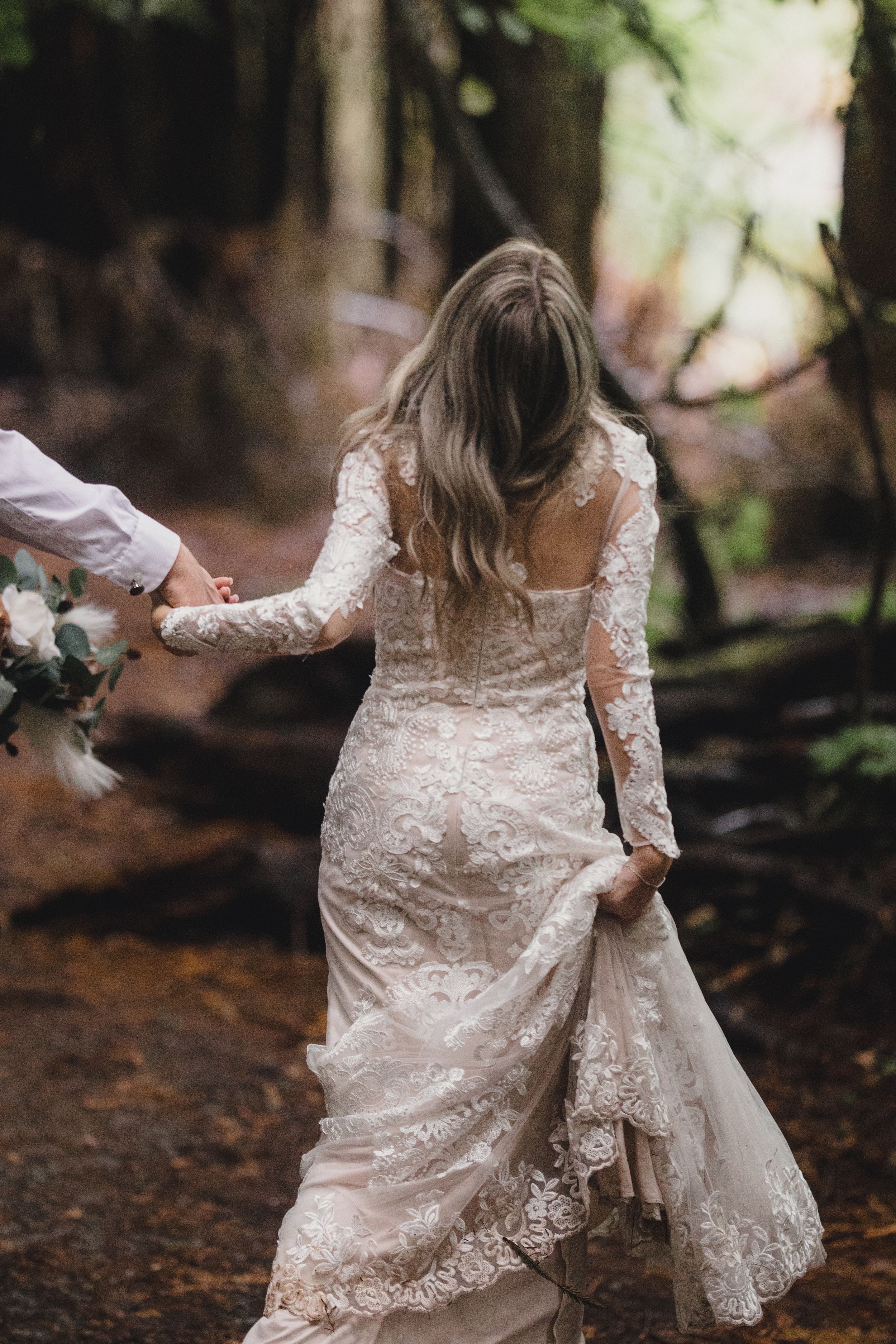 Bride in lace gown walks hand-in-hand through redwood forest on wedding day.