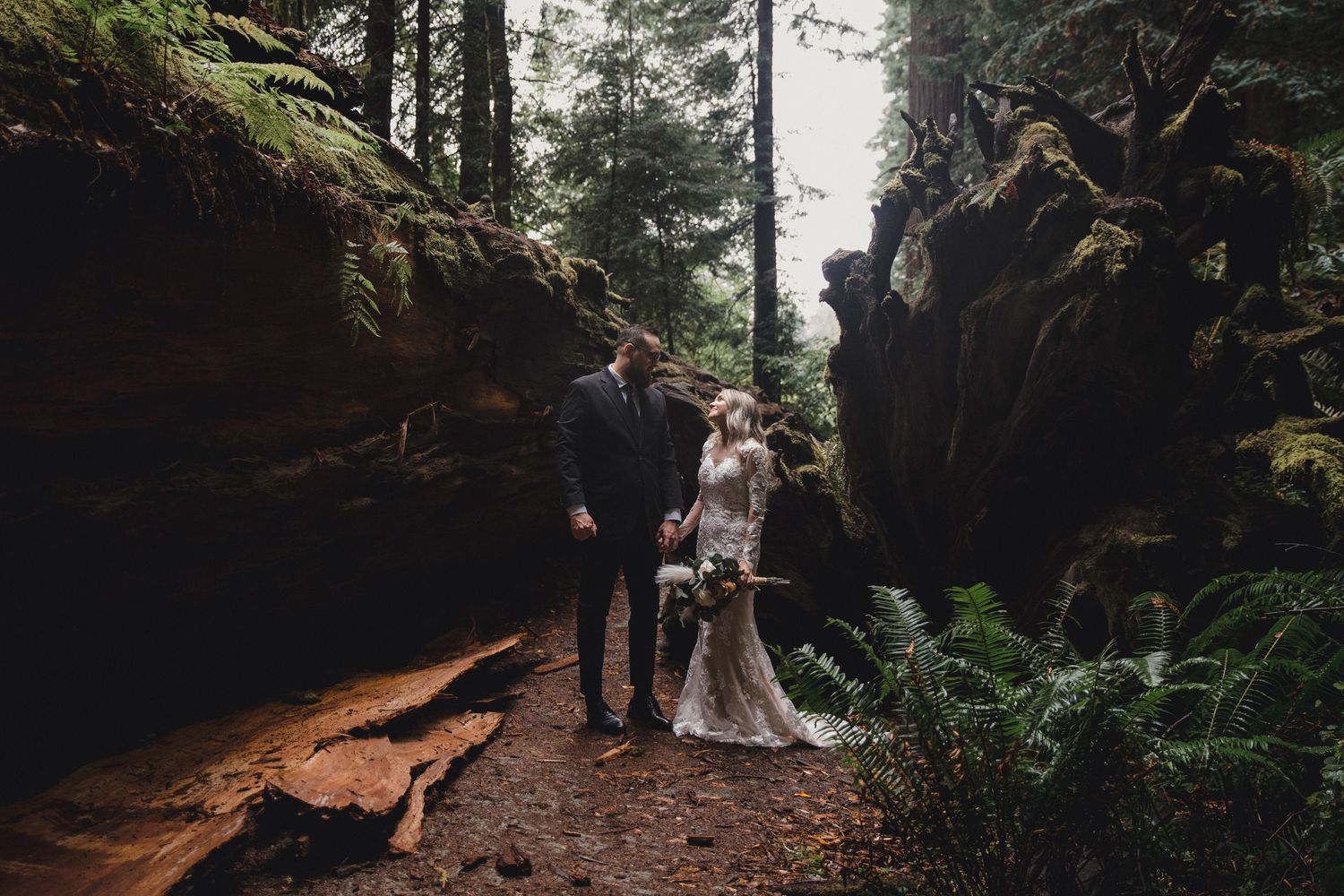 A couple stands beneath towering redwood trees during a romantic forest wedding ceremony.