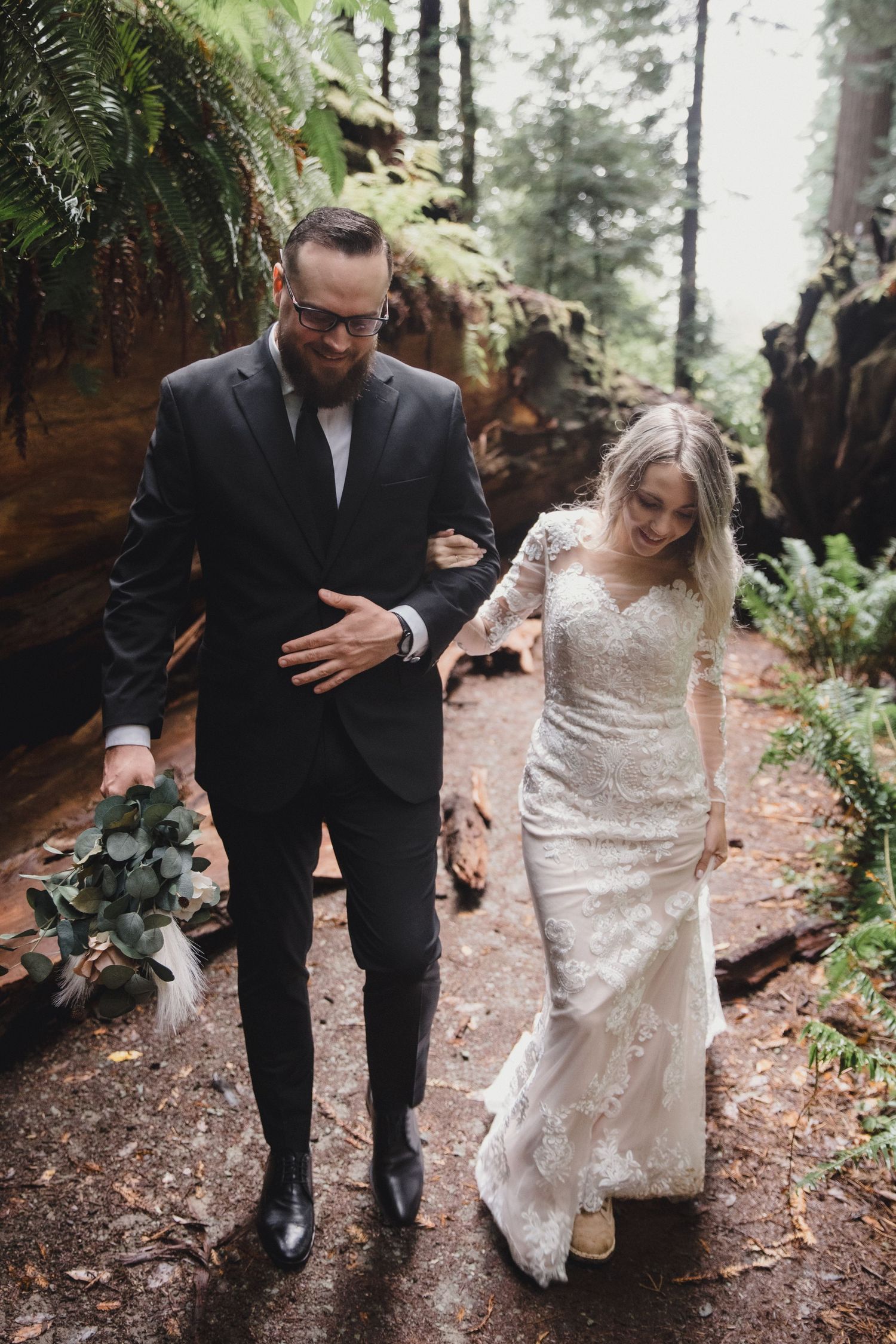 A couple walking together on a forest path, with the groom helping the bride in her white lace wedding dress.