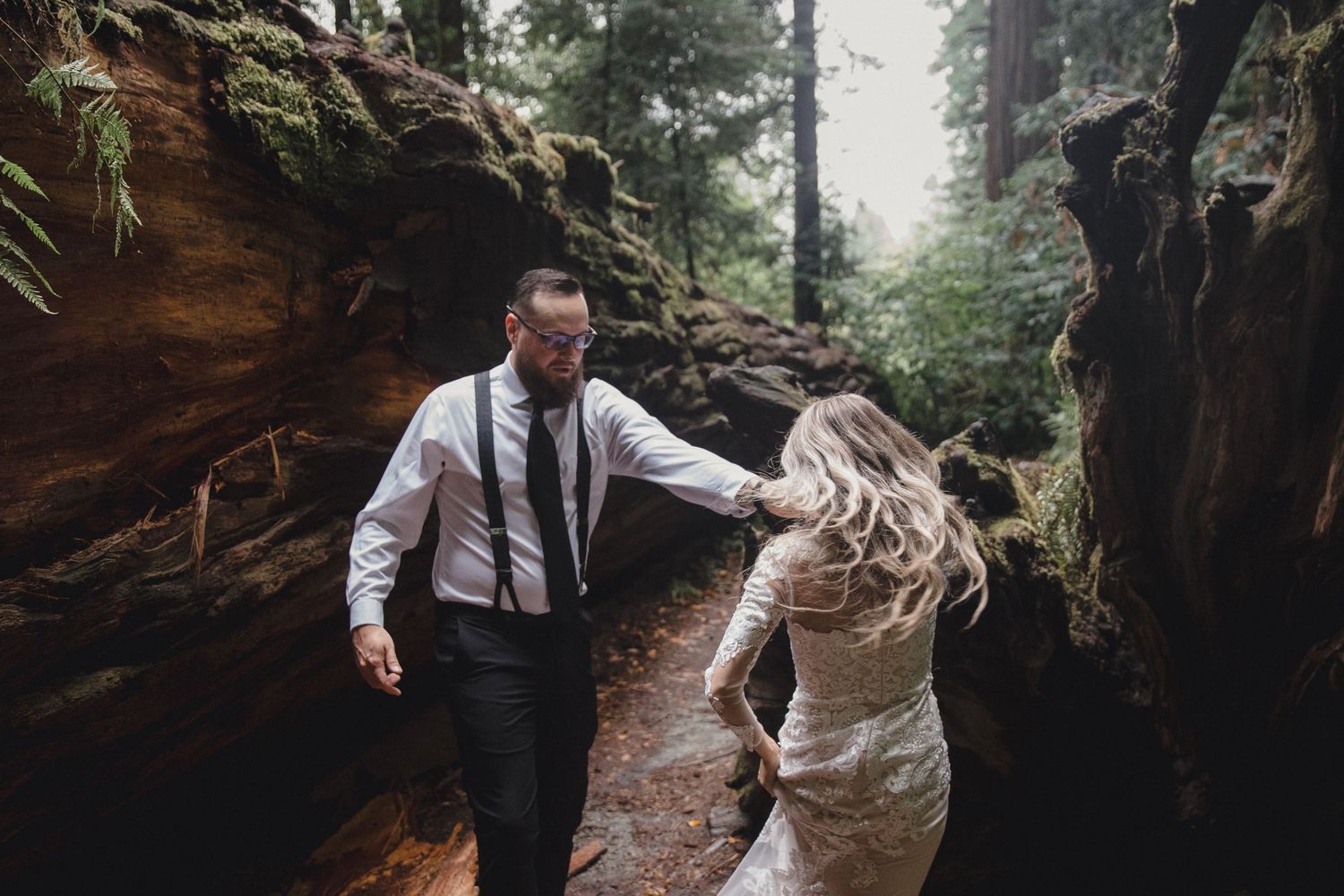 A couple in wedding attire walks hand in hand through a misty forest path between towering rock formations.