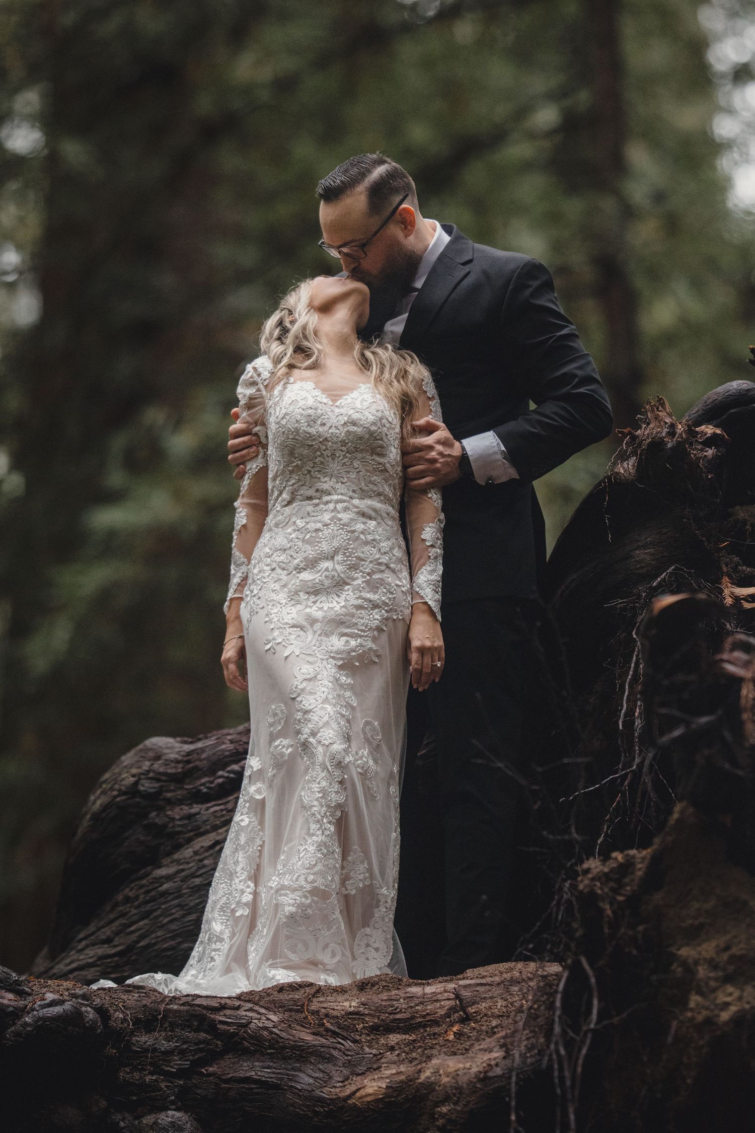 A couple in wedding attire poses dramatically on a fallen tree trunk in a moody forest setting.