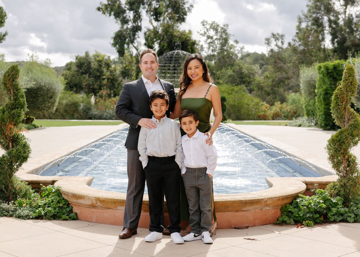 A family strolls together on a stone path next to a long reflecting pool in an elegant garden setting.