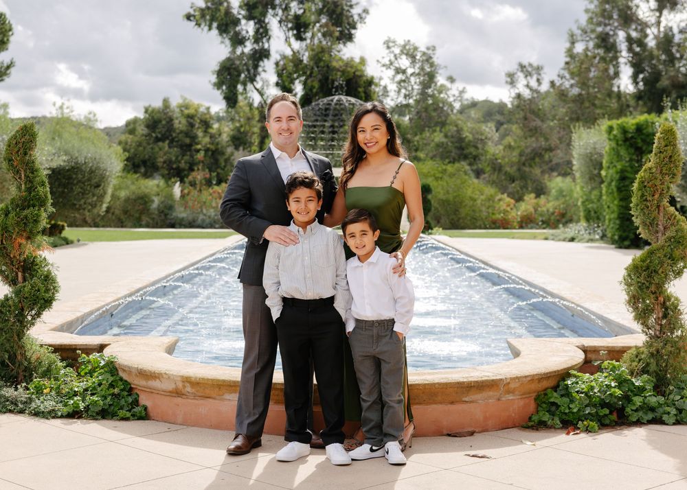 A family strolls together on a stone path next to a long reflecting pool in an elegant garden setting.