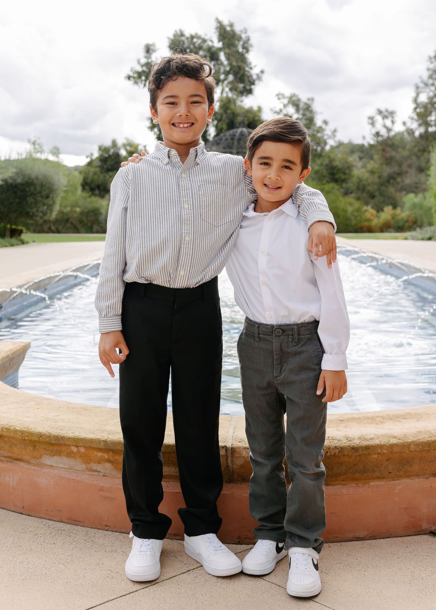 Two children in button-down shirts and casual pants pose together by a fountain on a sunny day.