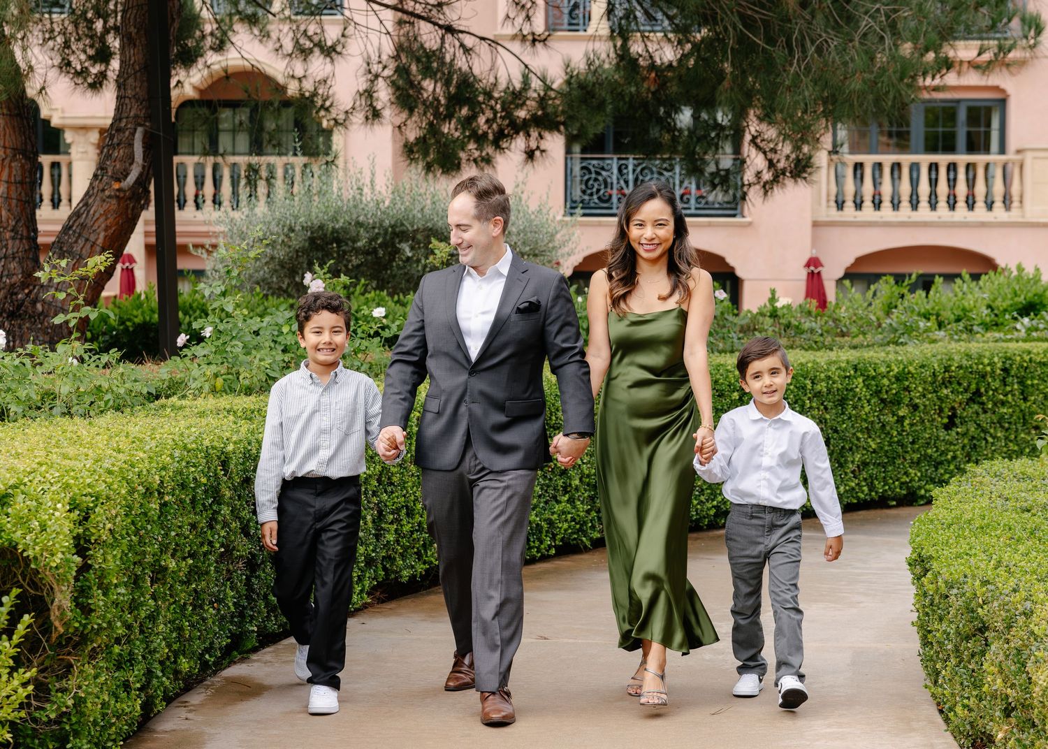 Family members walk together along a Mediterranean-style courtyard with pink walls and manicured hedges.