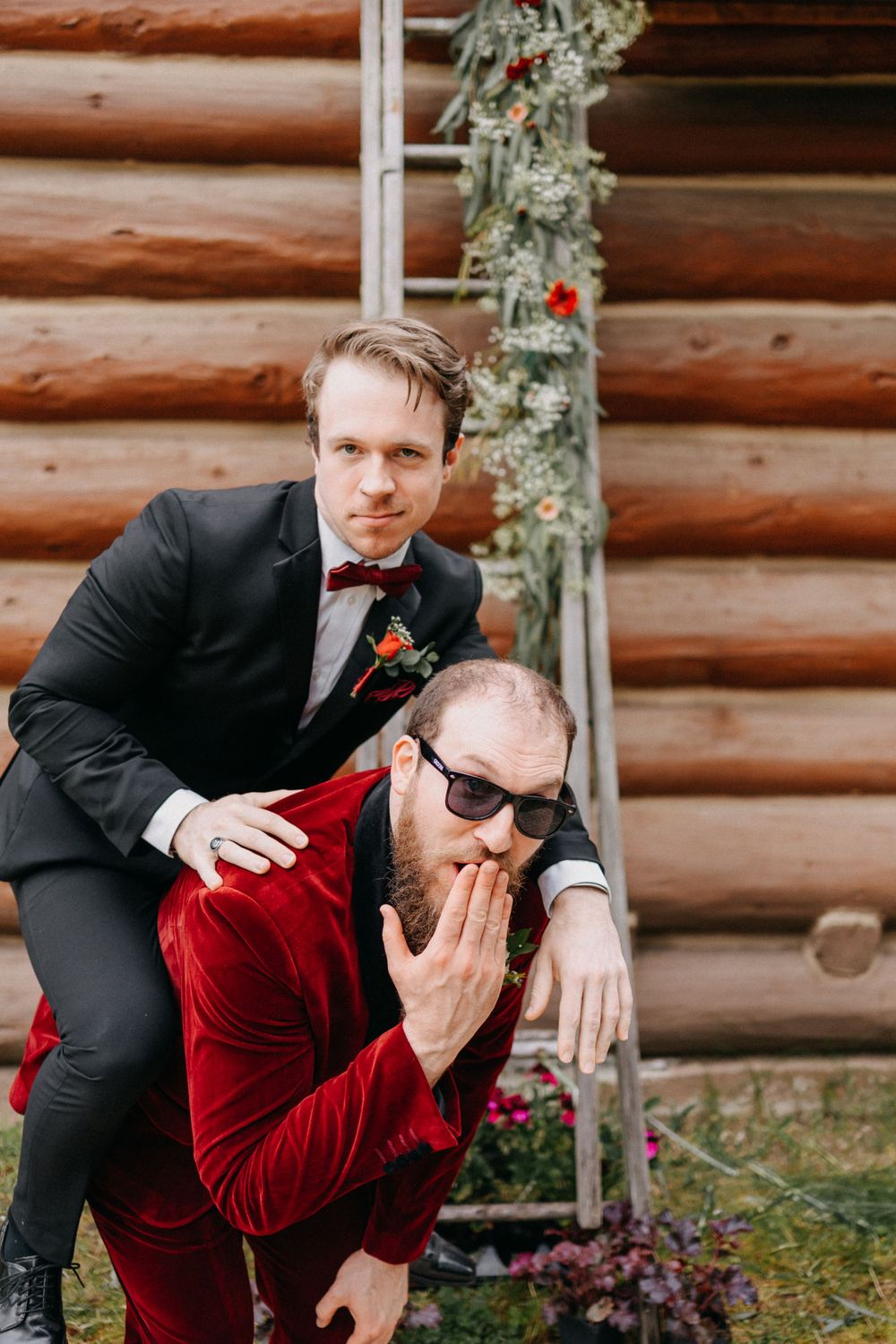 Two people in formal attire, one in a red velvet jacket, playfully posing near a rustic log cabin.