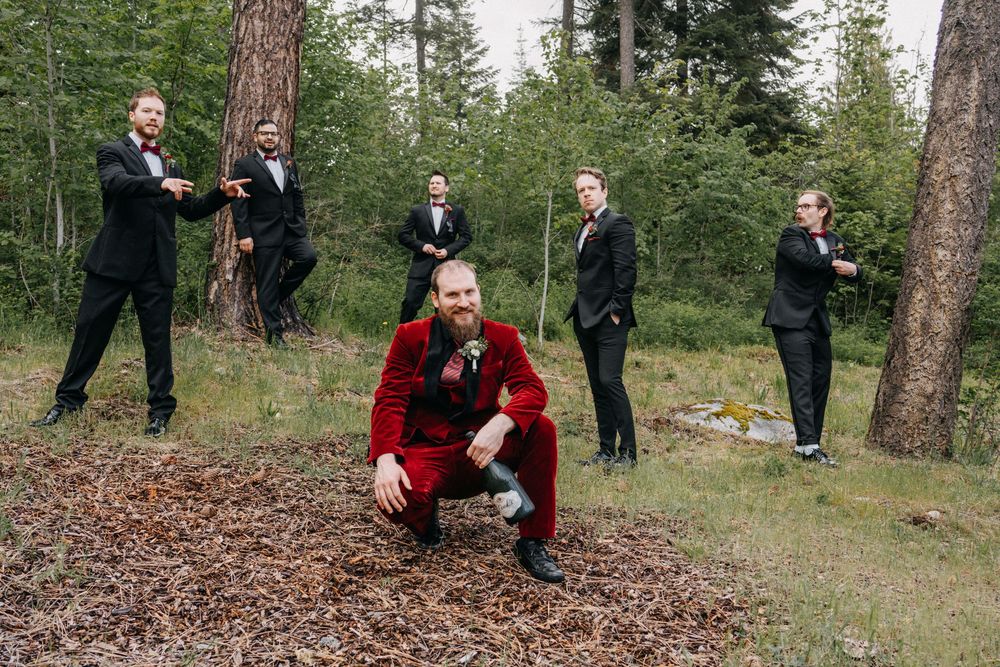 Wedding party members in burgundy and black pose playfully on the ground in a forest setting.