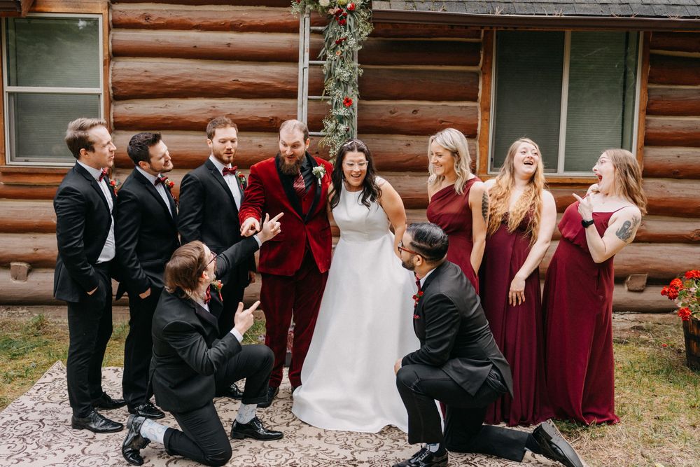 Wedding party in burgundy dresses and dark suits posing together in front of rustic log cabin exterior.