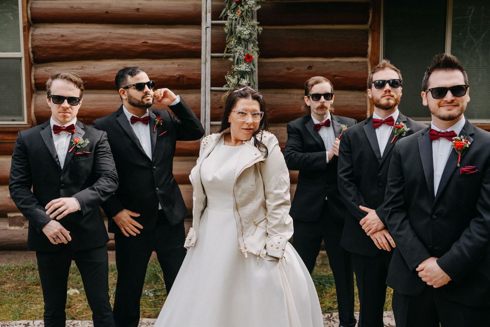 Wedding party lined up outdoors in formal attire with sunglasses against log cabin backdrop.