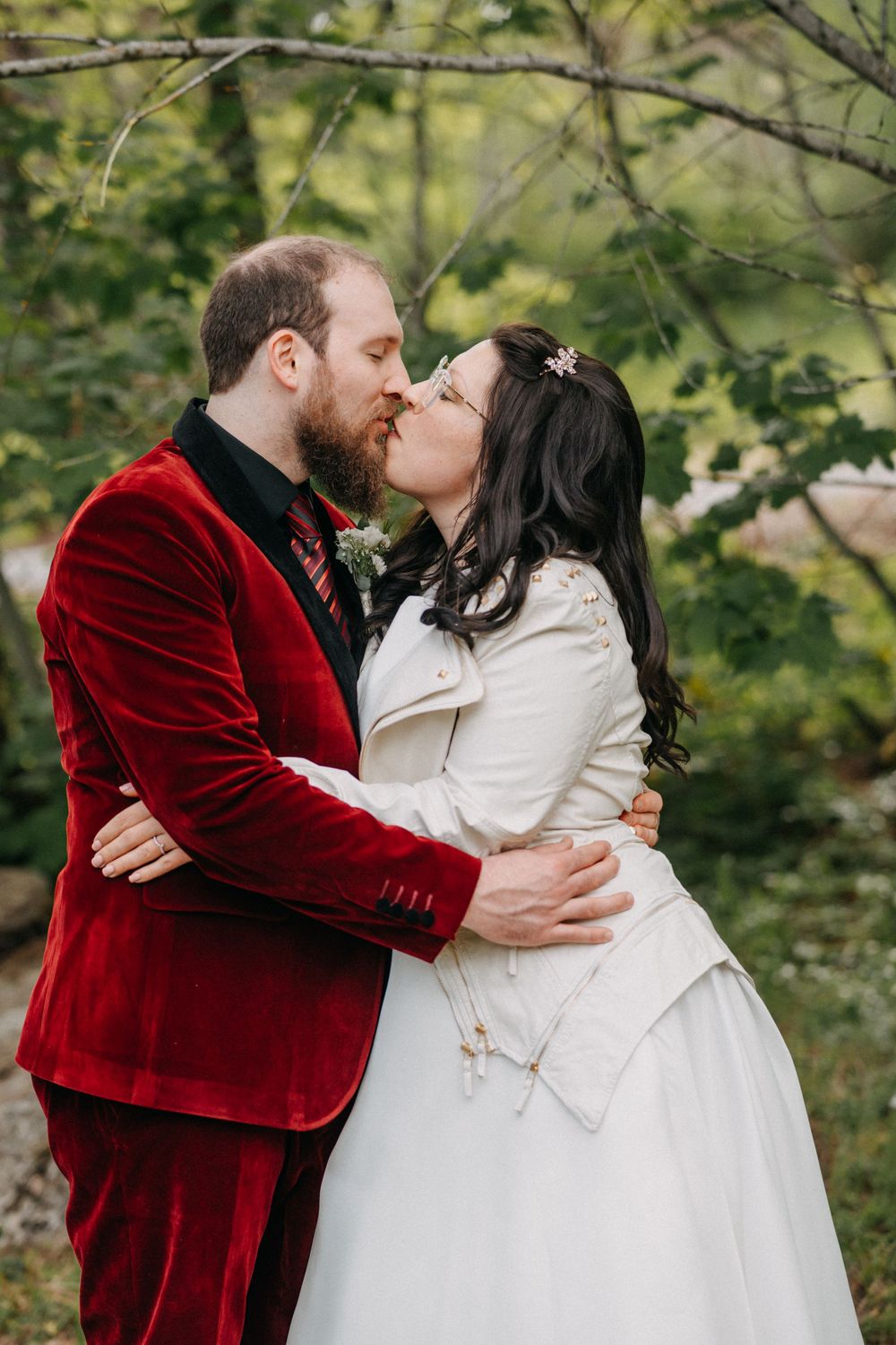 A romantic wedding couple embracing, with the groom in a deep red velvet jacket and bride in white.