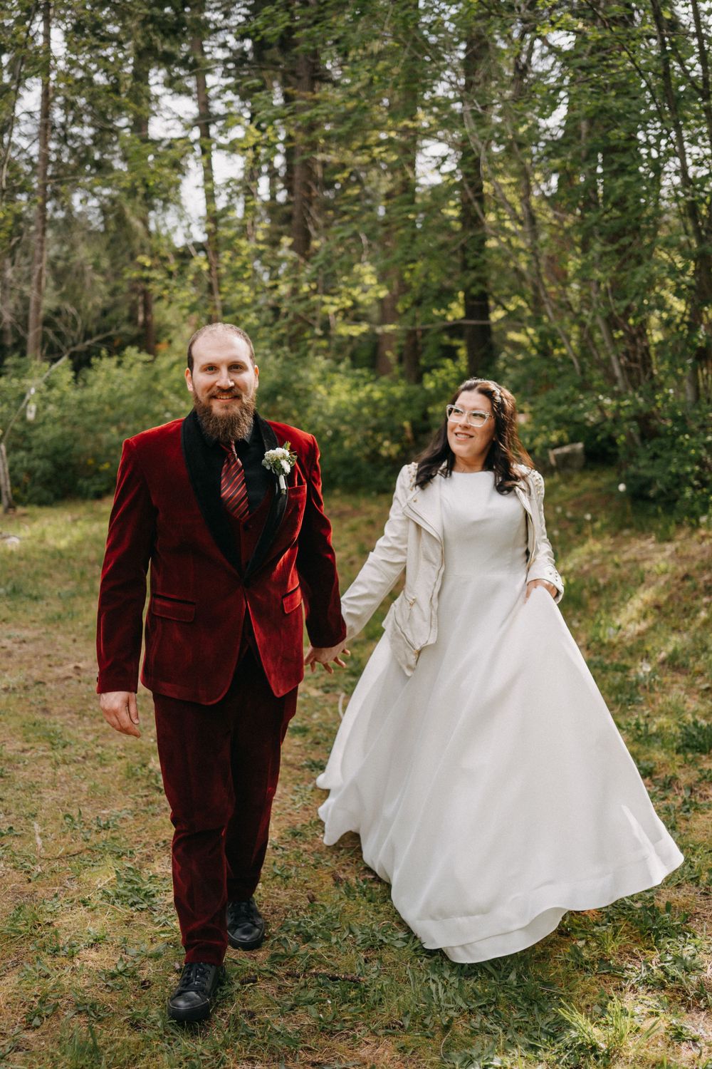 Couple in burgundy suit and white wedding dress walk together through wooded setting.