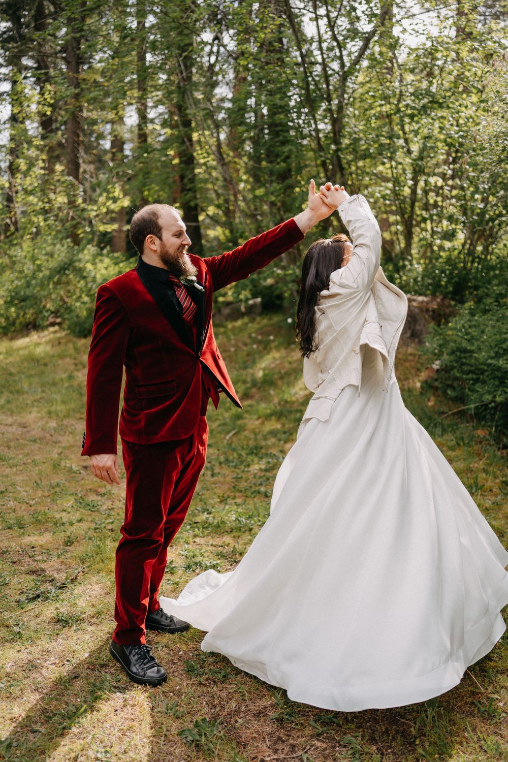 A couple in a burgundy velvet suit and flowing white wedding dress dance together in a sunlit forest setting.