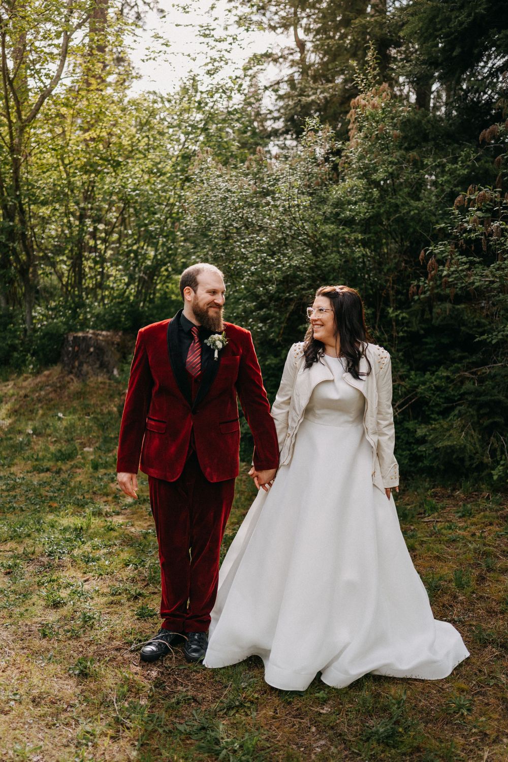 A couple in wedding attire walk together in a wooded setting, with the groom wearing a burgundy velvet suit.