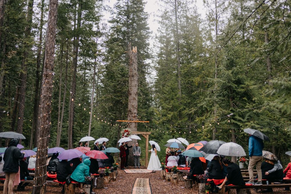 Outdoor forest wedding ceremony setup with colorful umbrellas and wooden cross altar.