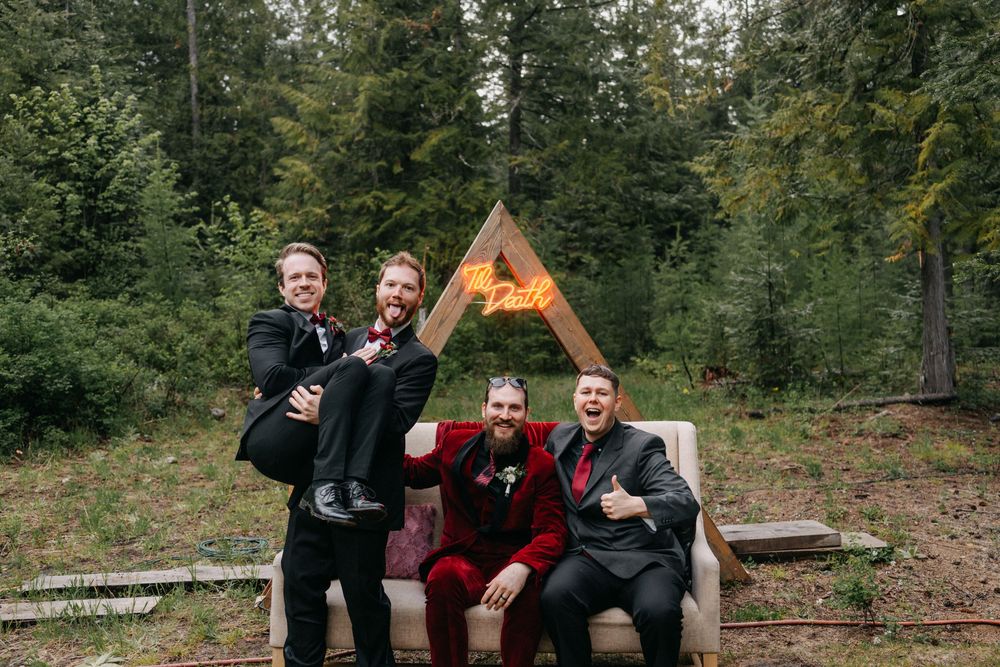 Wedding party members pose and celebrate together beneath a rustic wooden A-frame structure in the woods.