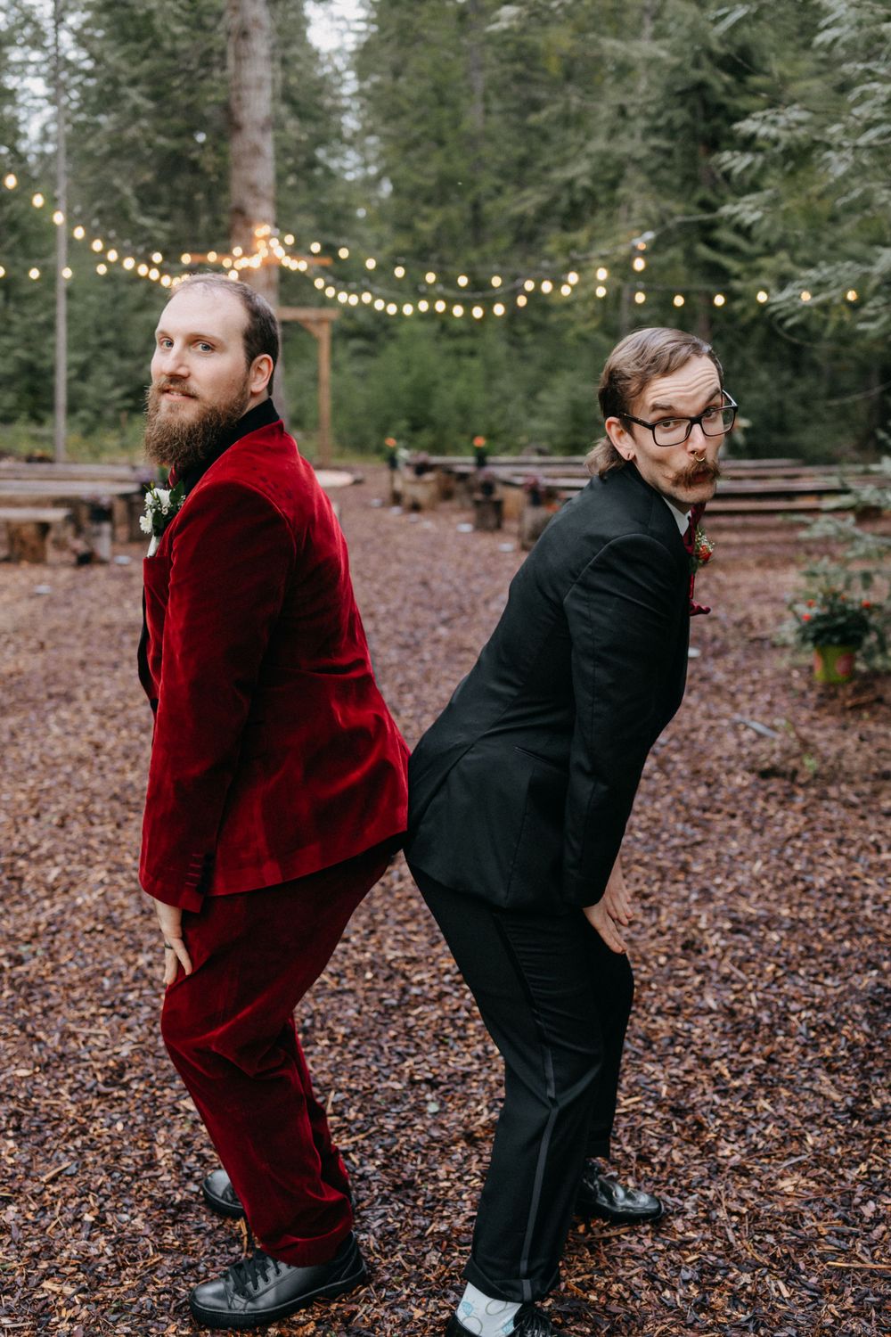 Two people in formal attire playfully pose in a forest setting with string lights overhead.