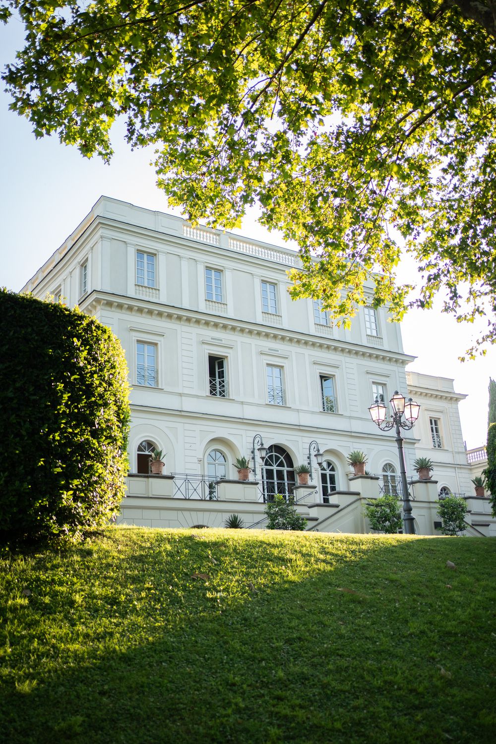Elegant white mansion with classical architecture viewed from sloping green lawn with overhanging tree branches.